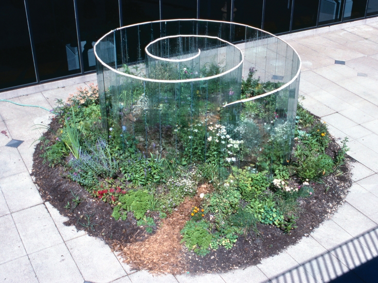 Installation view of Meg Webster's glass spiral, surrounded by growing plants and flowers.